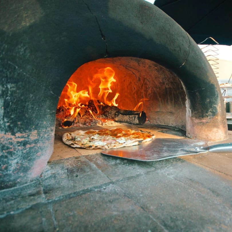 A pizza cooking inside a wood-fired oven with a metal peel, flames visible in the background.