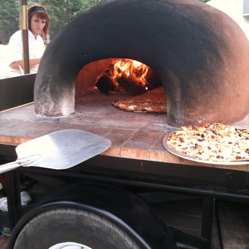 Versalia Pizza's mobile wood-fired oven with pizzas cooking inside and on a tray, a chef in the background.