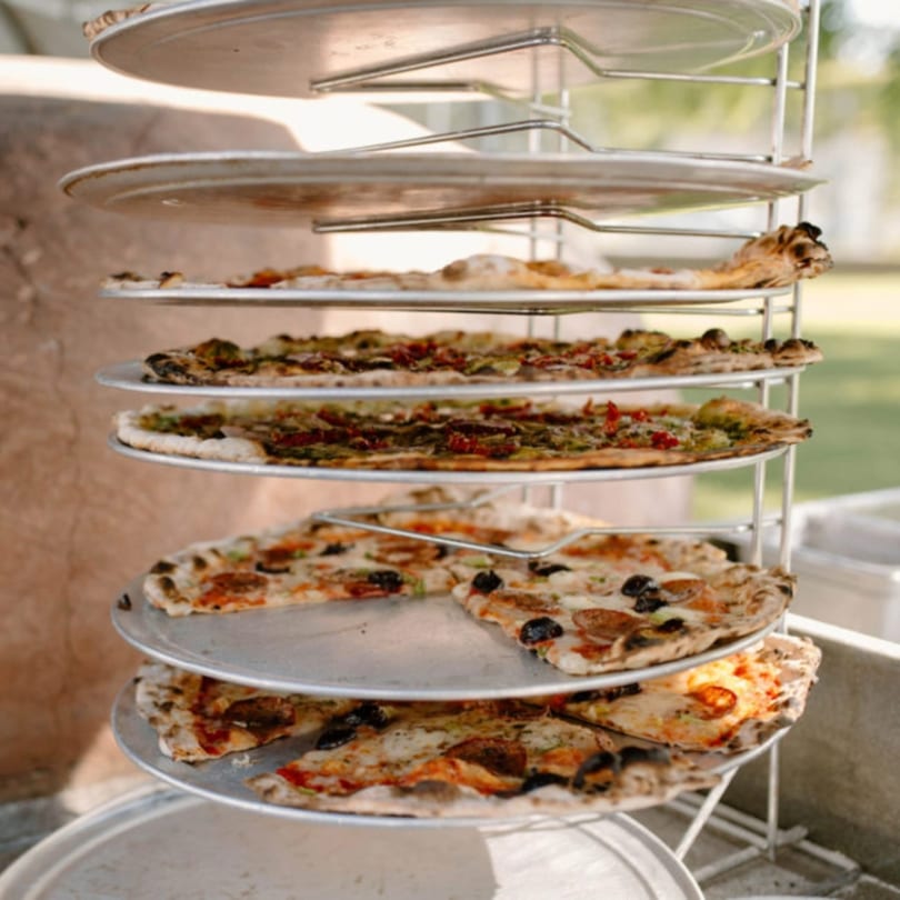 Multiple Versalia pizzas with various toppings are stacked on a metal rack outdoors.