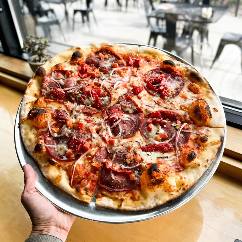 A hand holds a Versalia Pizza with pepperoni, red onions, and diced tomatoes, by a window.