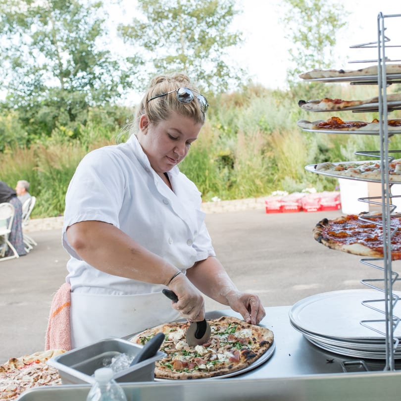 A Versalia Pizza chef slices a freshly baked pizza outdoors, with other pizzas on a rack nearby.