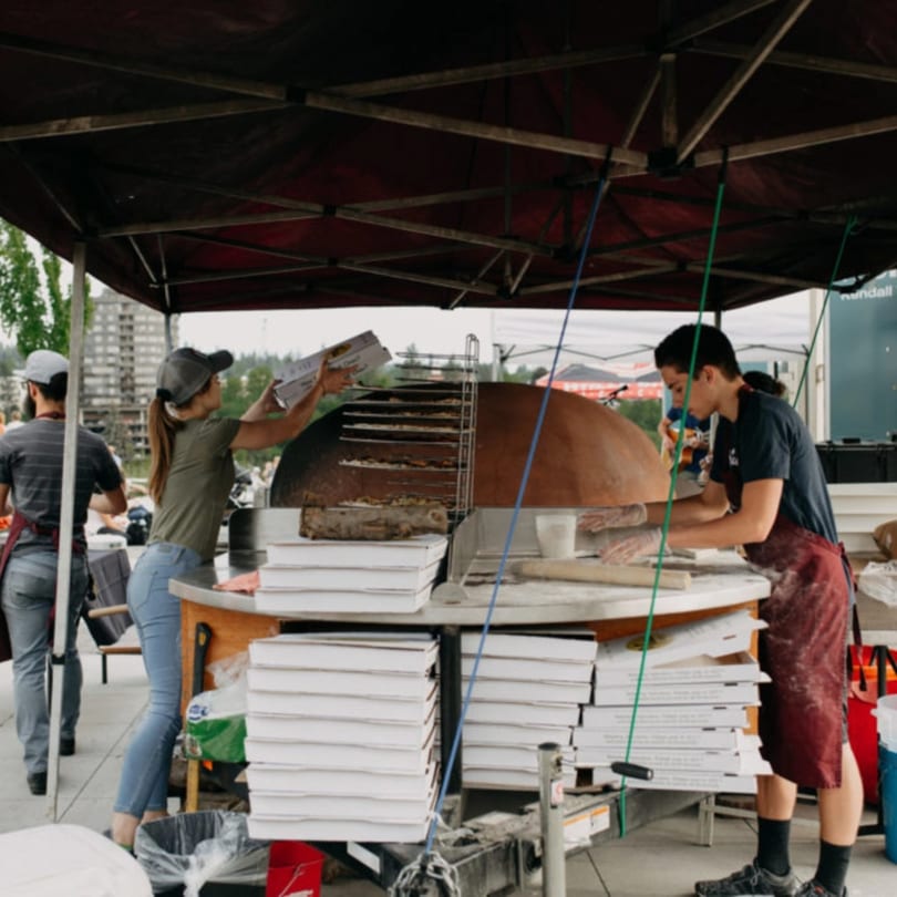 Versalia Pizza staff preparing pizzas at an outdoor event with a large wood-fired oven.