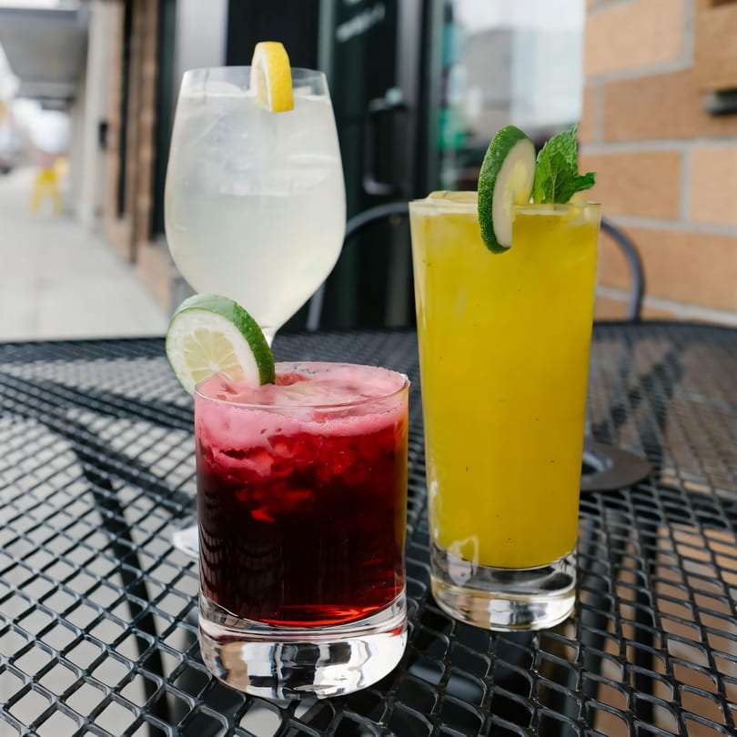 Three colorful cocktails, garnished with citrus and mint, sit on a black mesh outdoor table.