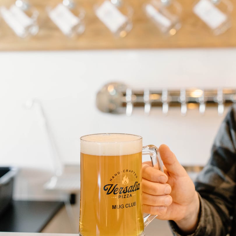 A hand holding a Versalia Pizza Mug Club glass filled with a foamy golden beverage.