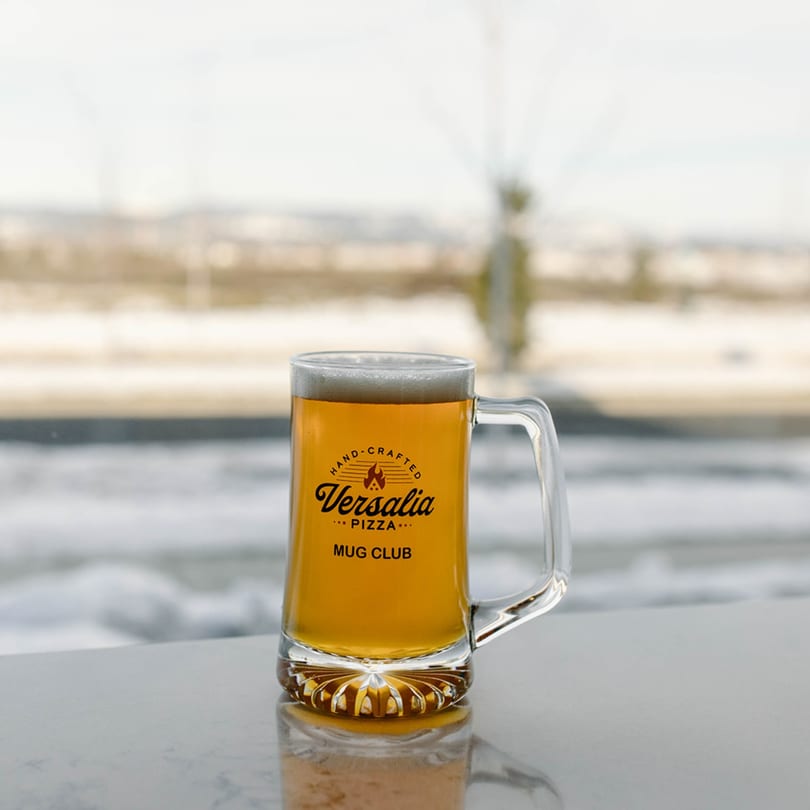 A Versalia Pizza Mug Club beer stein filled with golden beer and foam, against a snowy window.