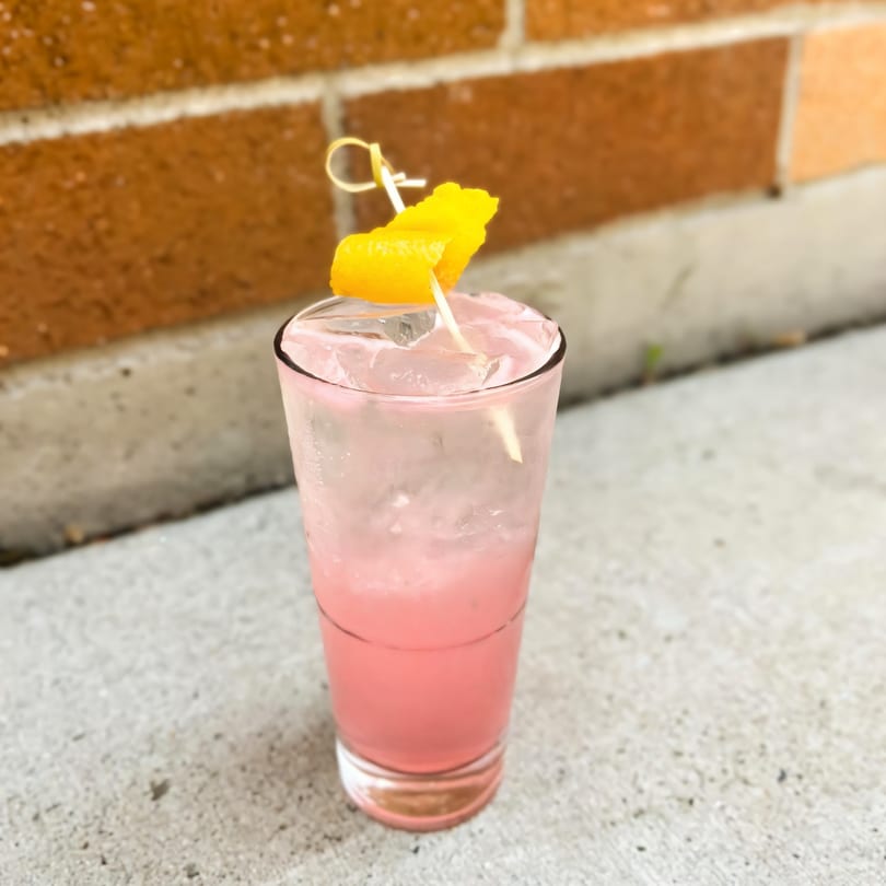 A tall glass of pink cocktail with ice and an orange peel garnish, against a brick wall.