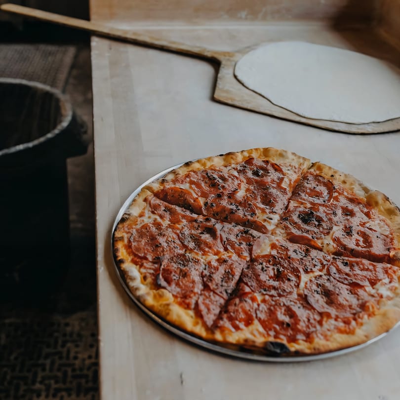 A freshly baked pepperoni pizza on a metal pan next to a pizza peel with raw dough.
